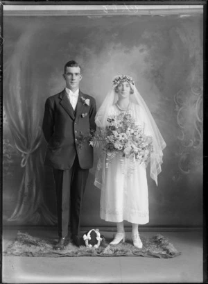 Studio portrait of unidentified wedding couple, groom in a stiff wing shirt collar, white bow tie and rose buttonhole, bride with long veil and pearl necklace holding flowers, with lucky horse shoe between them, Christchurch