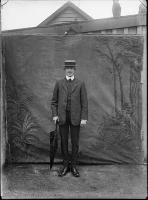 Outdoors portrait of unidentified man in a three pierce suit and straw hat with black band, standing with an umbrella in front of false backdrop, probably Christchurch region