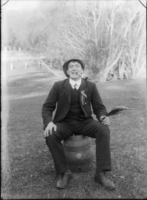 Outdoor portrait of unidentified man sitting on a beer keg in a three piece suit with lapel ribbons and double round shirt collar, striped tie and cloth cap, with a pipe holding a feather, probably Christchurch region