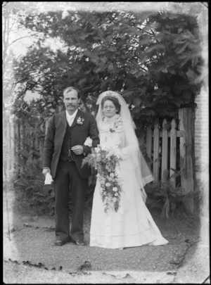 Outdoors unidentified wedding couple portrait on grass by a brick house, groom with large moustache and greenstone watch chain pendant, standing with bride with glasses and long veil holding flowers, probably Christchurch region