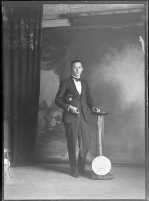 Studio portrait of an unidentified young male musician in a tuxedo and black bow tie holding a violin and bow, standing next to a wooden font with a tenor banjo in front, Christchurch