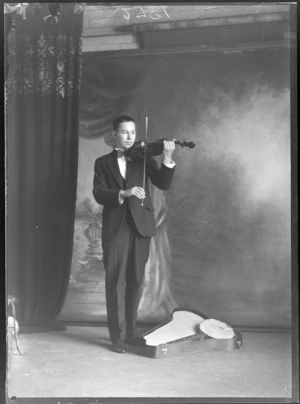 Studio portrait of an unidentified young male musician in a tuxedo and black bow tie standing playing a violin with a tenor banjo in front, Christchurch