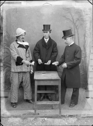 Outdoors in front of a false backdrop, three unidentified men in costume, a man in dog collar and large moustache and business man with top hats and cigars, with a third man in a clown's checked suit playing cards, probably Christchurch region