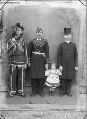 Outdoors in front of a false backdrop, three unidentified men and young girl in costume, a man in dog collar and large moustache and top hat, an American army officer and a native American Indian with a knife and and girl in [Pierrot?] costume, probably Christchurch region