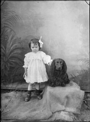 Outdoors in front of a false backdrop, portrait of a young unidentified girl standing on sacking in a lace dress with a large collar, with a black Cocker Spaniel dog, probably Christchurch region