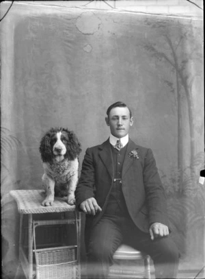 Studio portrait of an unidentified young man with double round shirt collar and three piece striped suit with lapel flower, sitting at a cane table with an old Cocker Spaniel dog, Christchurch