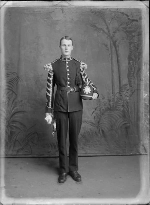 Studio portrait of an unidentified young [Lieutenant?] World War I soldier in full dress uniform, with sleeve and shoulder pads insignia, collar badges and bugle with tasselled cord, holding a spiked helmet with a large badge, Christchrch