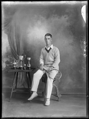 Studio unidentified portrait of a young adult male tennis player sitting holding his racket alongside a table with four cups of various sizes, Christchurch
