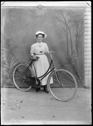 Outdoors in front of a false backdrop, portrait of a young unidentified woman in a lace dress with pearl necklace, heart shaped pendant, bar brooches, a metal belt and a hat, standing with a women's bicycle, probably Christchurch region