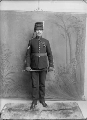 Studio portrait of an unidentified Sergeant World War I soldier in full dress uniform with handlebar moustache, sleeve insignia, collar badges and cord tie, a busby fur hat with plum, in leather groves holding a riding crop, Christchurch
