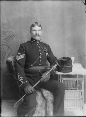 Studio portrait of an unidentified Sergeant World War I soldier in full dress uniform, with handlebar moustache, sleeve insignia [artillery?], collar badges and cord tie, in leather groves holding a riding crop sitting with busby fur hat with plum on a table, Christchurch