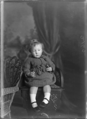 Studio portrait of unidentified young girl in a knitted woollen dress and leather shoes with pom poms sitting on a wooden high chair, Christchurch