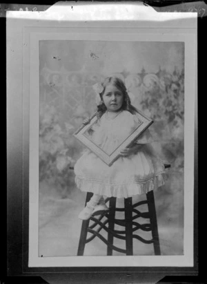 Studio portrait of a young girl [Miss E Fox?] in a lace dress, pearl necklace, hair curls and ribbon, sitting on a high stool holding a picture frame around her neck, location unknown.