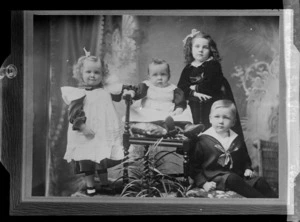 Studio family portrait of four young children, two girls in lace dresses and hair bows, and a boy in a sailor suit gathered around a baby in a lace pinafore sitting on a wooden chair, location unknown.