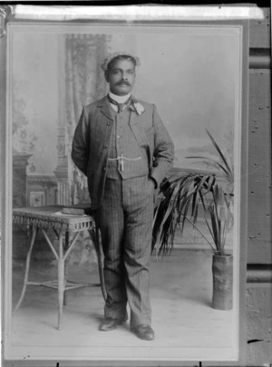 Studio portrait of a man with moustache, in a striped three piece suit with lapel flower, cloth cap and greenstone watch chain pendant, standing in front of a table with a book,location unknown.