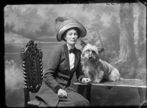 Studio portrait of an older woman [Mrs Richmond?], in a jacket, high collar shirt, dark bow tie and large hat, sitting at a table with a small dog, Christchurch