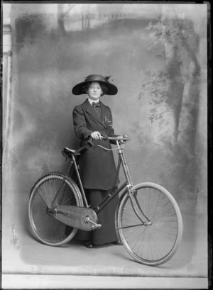Studio unidentified portrait of a young woman in a double round shirt collar with bow tie, a flower patterned blouse, matching coat and long skirt, with a large feathered hat with hat pins, standing with a women's bicycle, Christchurch