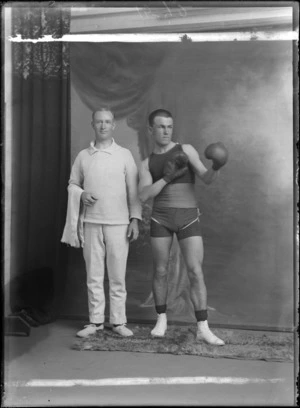 Studio portrait of two unidentified men, one wearing a boxing outfit with shorts, boots and boxing gloves, the other wearing a jersey and pants and holding a towel, most likely boxer and coach, Christchurch