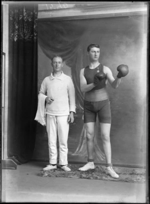 Studio portrait of two unidentified men, one wearing a boxing outfit with a medal pinned on it [war medal?], shorts, boots and boxing gloves, the other wearing a jersey and pants and holding a towel, most likely boxer and coach, Christchurch