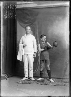 Studio portrait of unidentified man and boy, one wearing a boxing outfit with shorts, boots and boxing gloves, the other wearing a jersey and pants and holding a towel, most likely boxer and coach, Christchurch