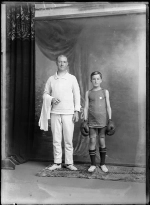 Studio portrait of unidentified man and boy, one wearing a boxing outfit with shorts, boots and boxing gloves, the other wearing a jersey and pants and holding a towel, most likely boxer and coach, Christchurch