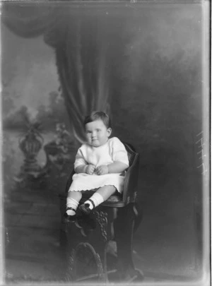 Studio portrait of an unidentified baby sitting in a highchair wearing an embroidered gown, socks and leather booties, Christchurch