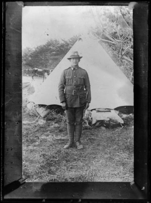 Unidentified soldier in uniform including, collar and hat badges, belt and bayonet, standing by a tent with horses in the background, [mounted rifles?]