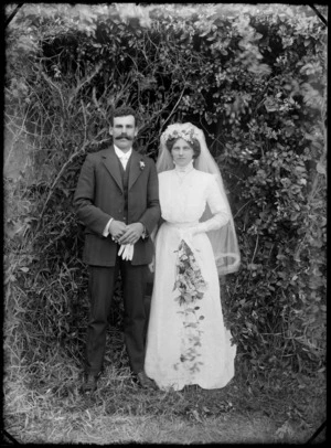 Outdoor portrait of unidentified wedding couple in front of trees, groom with walrus moustache and bride with long veil and cross necklace holding flowers, probably Christchurch region