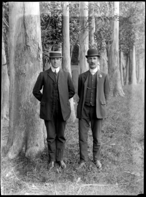 Outdoors portrait of two unidentified men in front of gum trees, older man with moustache, buttonhole and bowler hat, the younger man in pinstriped suit and straw hat, both with double round shirt collars, probably Christchurch region