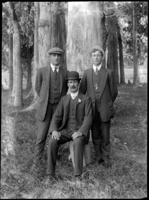 Outdoors portrait of three unidentified men in three piece suits and double round shirt collars and hats, with greenstone tiki and medallion watch chain pendants, probably Christchurch region