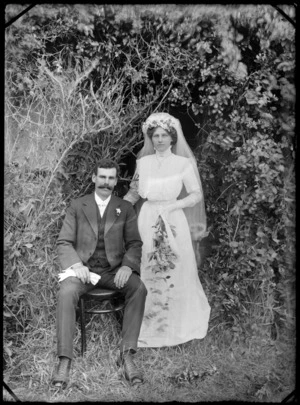 Outdoor portrait of unidentified wedding couple in front of trees, groom with walrus moustache sitting, bride standing with long veil and cross necklace holding flowers, probably Christchurch region