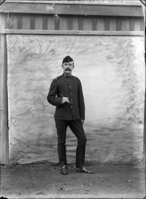 Outdoors portrait of unidentified World War I soldier with large moustache, collar and shoulder badges, forage cap and stirrups holding a riding crop [Mounted Rifles?], taken in front of false backdrop, probably Christchurch region