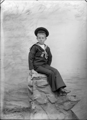 Outdoors portrait of unidentified young boy in a sailors' uniform sitting on sacks, with braided whistle cord and cap with 'HMS Nautilus', taken in front of false backdrop, probably Christchurch region