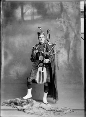 Studio portrait of unidentified World War I soldier in full dress highland piper uniform, with doublet jacket and 'CCS' sporran, tartan kilt and cape with shoulder and belt buckles, bagpipes and cap battalion badge with feather, Christchurch