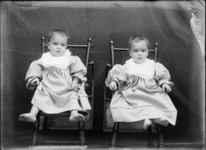 Outdoors portrait of unidentified twin babies with large lace bibs sitting on small wooden chairs, taken in front of a false blanket backdrop, probably Christchurch region