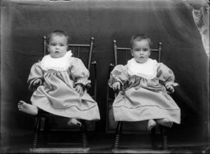 Outdoors portrait of unidentified twin babies with large lace bibs sitting on small wooden chairs, taken in front of a false blanket backdrop, probably Christchurch region