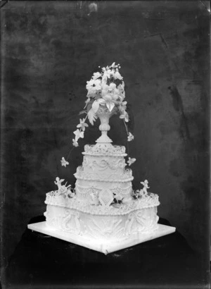 Studio portrait of a three tiered wedding cake with a 'AMR EJS' sign, cherubs, decorative icing and a vase with flowers on top, Christchurch