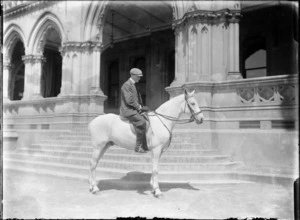 Exterior portrait outside building of unidentified man on horse, wearing a cap