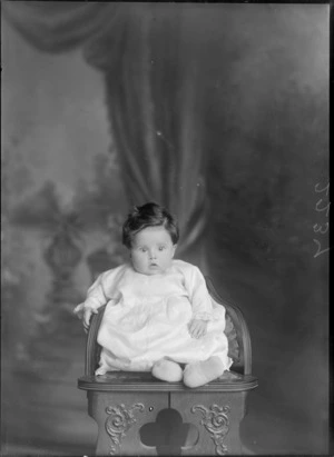 Studio portrait of an unidentified baby in a christening gown and woollen socks sitting on a wooden highchair, Christchurch