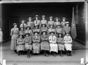 Outdoors portrait of the Sydenham Girl Scouts Troop with twenty one unidentified young women and one adult den mother troop leader with merit badges and company flag, taken in front of a large wooden building loading bay, Sydenham, Christchurch