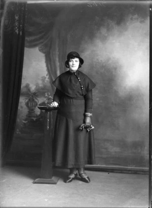 Studio portrait of unidentified woman with bobbed haircut wearing hat, cape with large buttons, dress, leather gloves, high heeled shoes, Christchurch