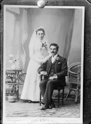 Studio unidentified wedding couple portrait, groom with moustache, greenstone watch chain pendant and lapel flowers sitting, bride with long veil standing, Photograph by Wrigglesworth and Binns, Christchurch or Wellington