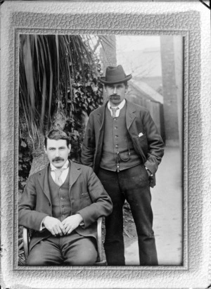 Outdoors in front of a cabbage tree with brick buildings beyond, two unidentified young men with moustaches and three piece suits, one standing with greenstone watch chain pendant, patterned tie and hat, location unknown