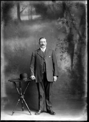 Studio unidentified portrait of a man with handlebar moustache, Arundel shirt collar and patterned tie, striped three piece suit and watch chain pendant, standing with bowler hat on a table, Christchurch
