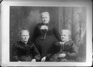Studio unidentified portrait of three older women [sisters?] two of which [twins?], in dark lace dresses with bar brooches, a cravat and large lace collar, one standing with two sitting, location unknown