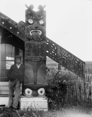 Unidentified young Maori man standing alongside a carving on the front gable of Kikopiri meeting house in the Horowhenua
