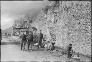 New Zealanders stroll pass women washing clothes in a stream near ancient wall in Alife in Italy, World War II - Photograph taken by George Kaye