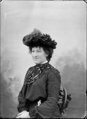 Studio portrait of unidentified woman in dark cotton blouse with lace and sequins and dark hat sitting in a chair, Christchurch