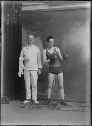 Studio portrait of two unidentified men, one wearing a boxing outfit with shorts, boots and boxing gloves, the other wearing a jersey and pants and holding a towel, most likely boxer and coach, Christchurch