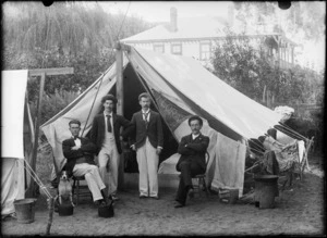 Outdoors portrait of four unidentified young men with moustaches in front of 'Flora' tent with a dog and 'Aulsebrook Biscuits' tin and other camping gear, probably Christchurch region
